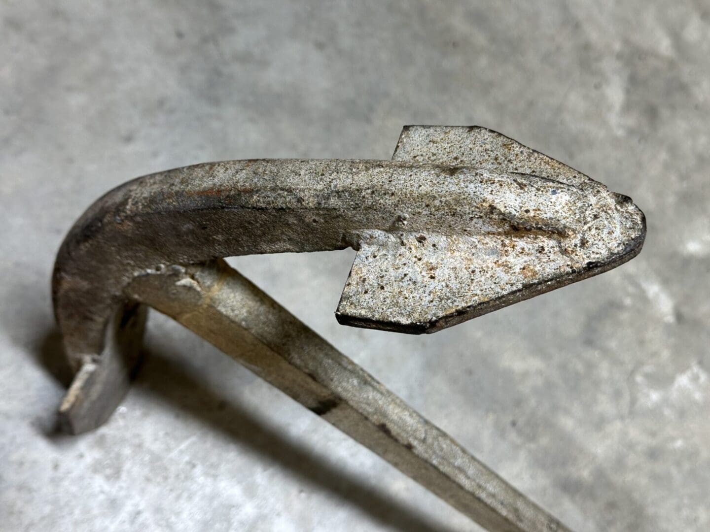 Close-up of a rusty, bent metal hook with a pointed end on a concrete floor.
